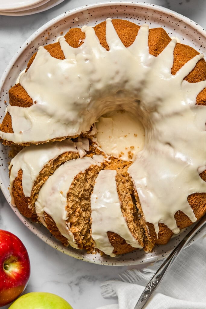 A sliced vegan gluten-free apple cake with white icing rests on a speckled plate, shown next to red and green apples on a marble surface. A serving utensil and napkin are also visible beside the cake.