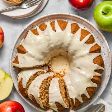 A glazed vegan gluten-free apple cake with several slices cut, surrounded by red and green apples on a marble surface. A plate with a cake slice and two forks is in the corner. A pie server rests nearby.