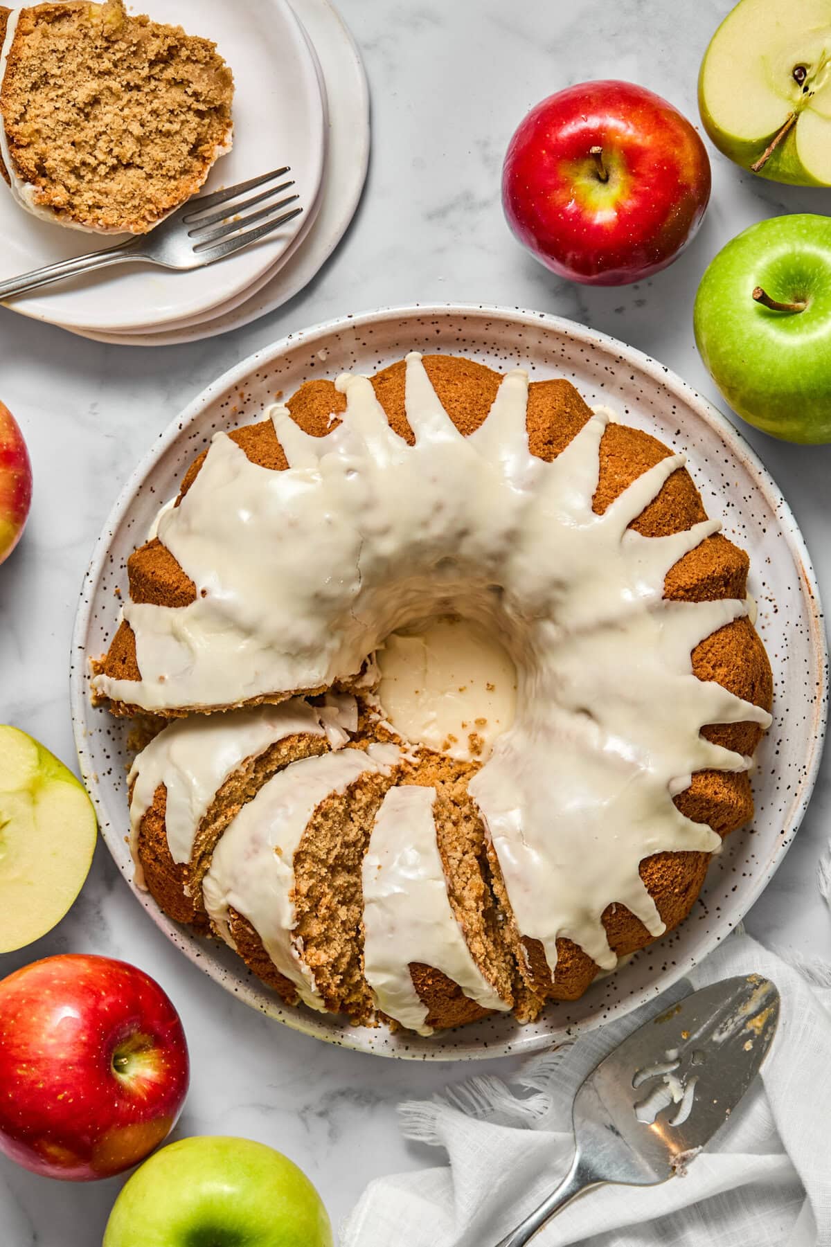 A glazed vegan gluten-free apple cake with several slices cut, surrounded by red and green apples on a marble surface. A plate with a cake slice and two forks is in the corner. A pie server rests nearby.