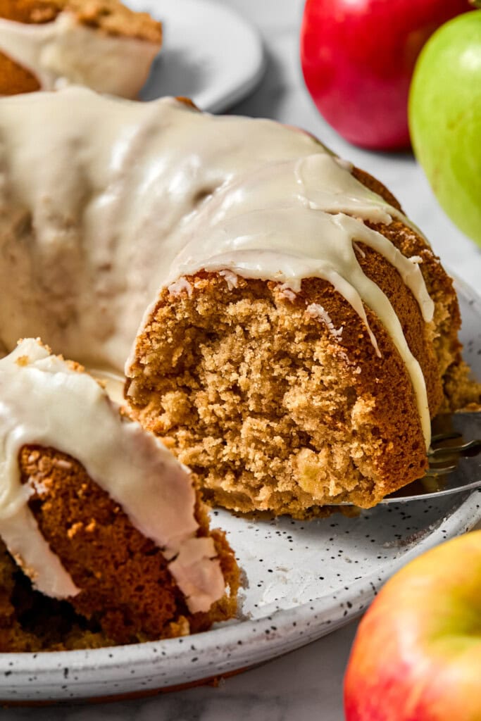 A sliced vegan gluten-free apple cake with light brown icing sits on a speckled plate, surrounded by red and green apples. The cake has a moist, dense texture and a slice is being served.