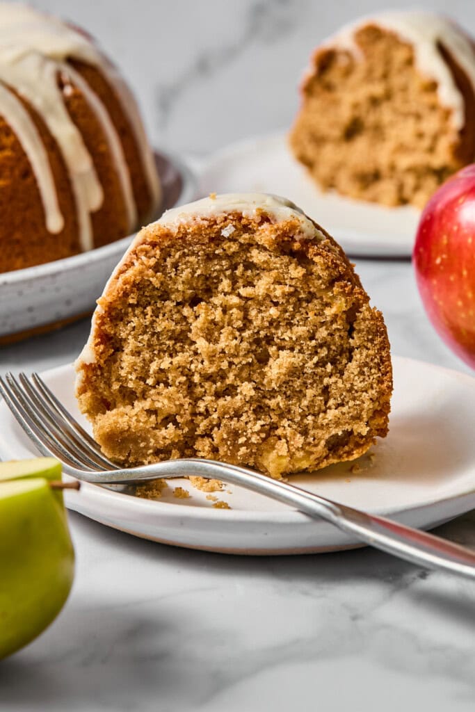 A slice of spiced vegan gluten-free apple cake with white icing sits on a plate with a fork. An apple and part of a green apple are nearby, and more cake is in the background.