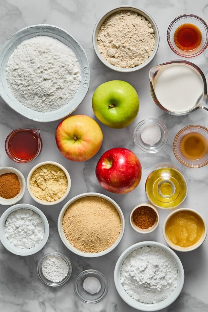 Top-down view of various baking ingredients in bowls and jars—flour, sugar, apples, spices, milk, honey, and applesauce—arranged neatly on a white marble surface, ready for making a delicious vegan gluten-free apple cake.