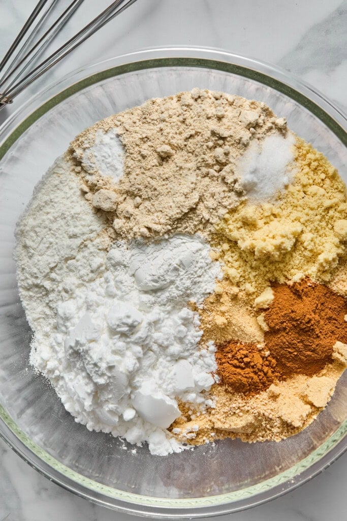 A glass bowl containing separate piles of flour, almond flour, ground spices, baking powder, baking soda, and salt on a marble countertop—perfectly prepped ingredients for making a vegan gluten-free apple cake—with metal whisks nearby.