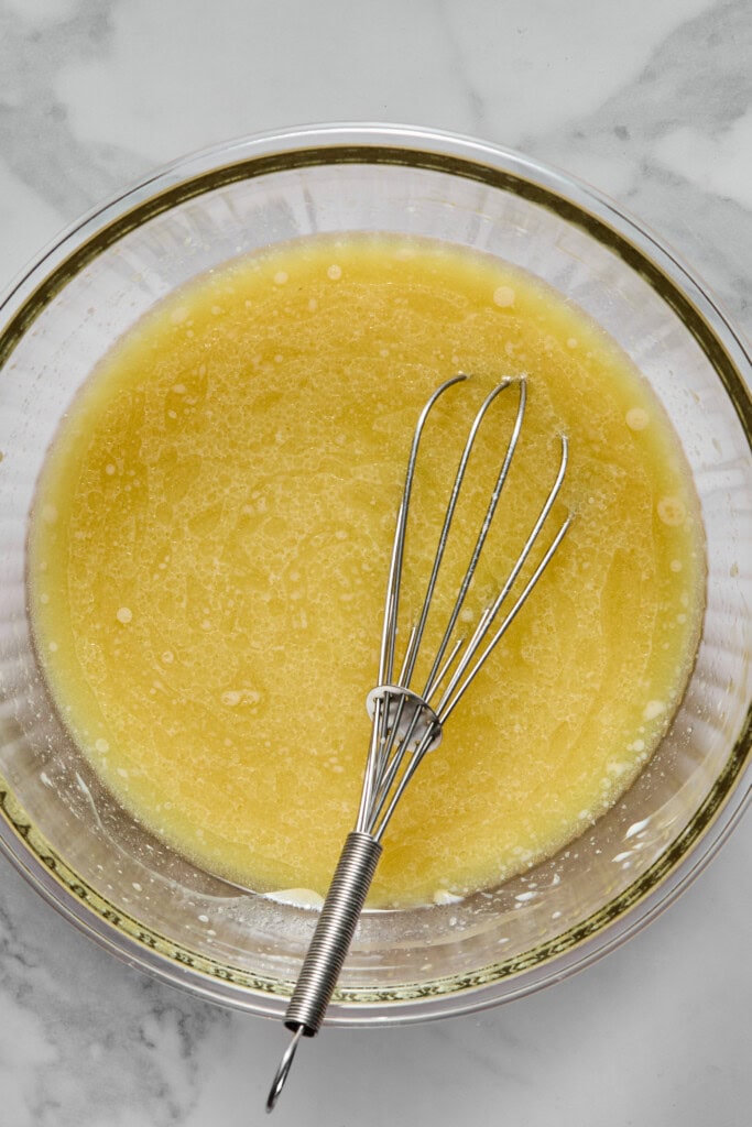 A metal whisk rests in a glass bowl filled with a yellow liquid mixture on a marble surface, ready for preparing a vegan gluten-free apple cake.
