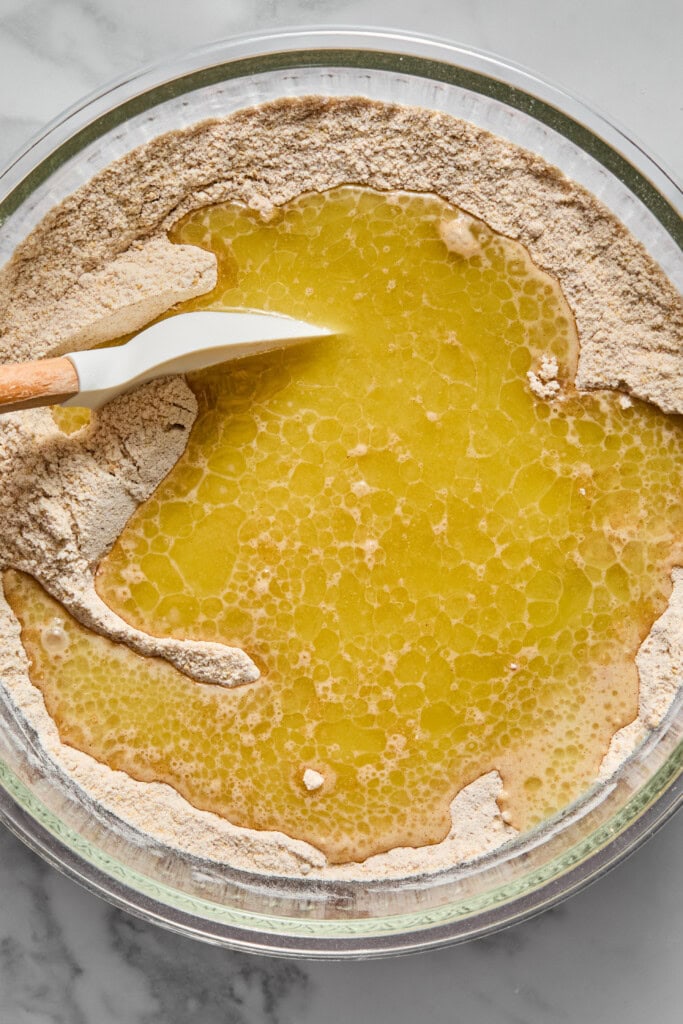 A glass bowl with flour and melted butter being mixed with a spatula for a vegan gluten-free apple cake, ready for baking, on a marble countertop.