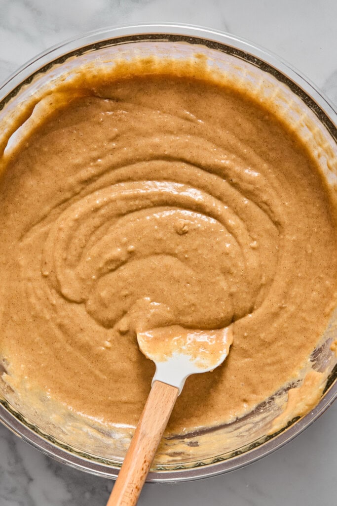 A glass bowl filled with light brown vegan gluten-free apple cake batter is being mixed with a white spatula on a marble countertop.