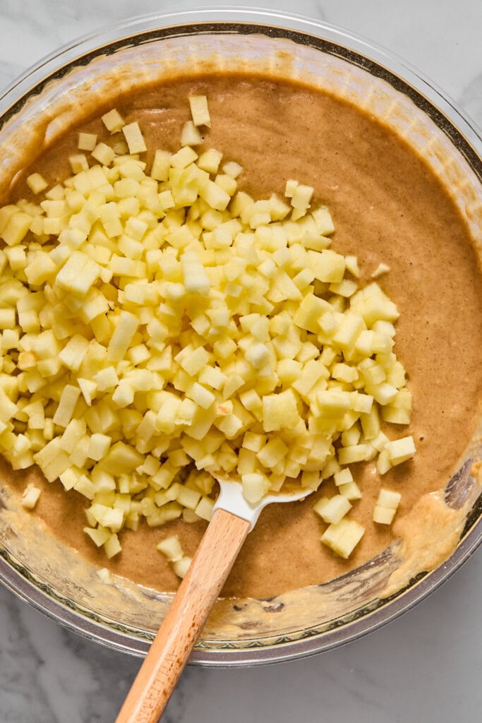 A glass bowl filled with batter for a vegan gluten-free apple cake, topped with a pile of small, diced apples. A spatula with a wooden handle rests in the mixture, ready for mixing. The bowl sits on a light marble surface.