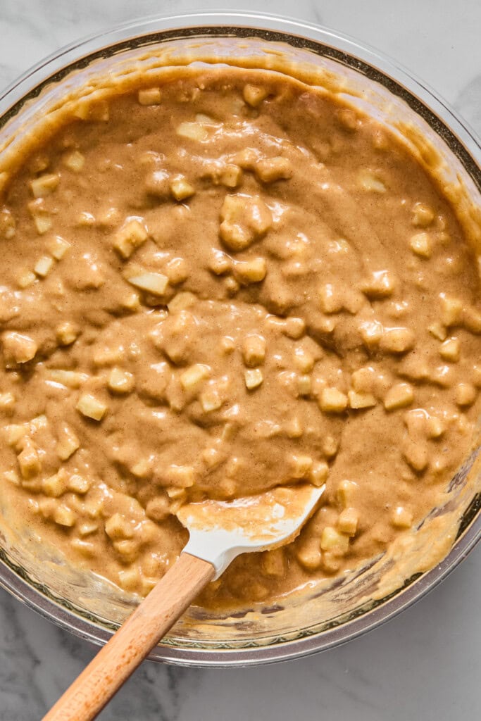 A glass bowl filled with chunky vegan gluten-free apple cake batter, being mixed with a white spatula on a marble counter.