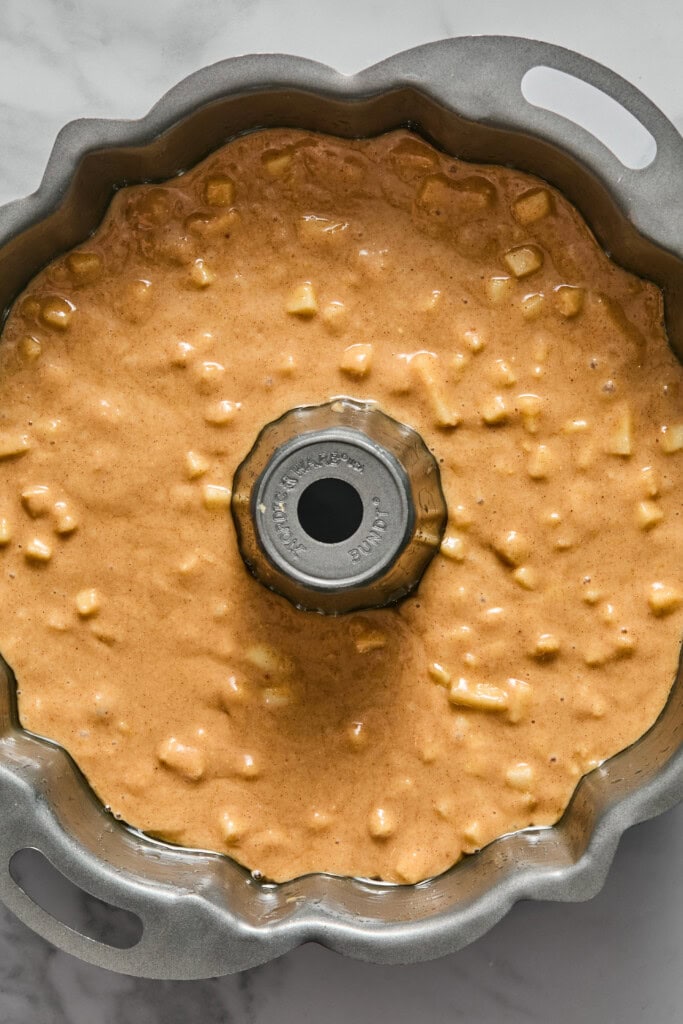 A bundt pan filled with unbaked vegan gluten-free apple cake batter containing small chunks, viewed from above on a marble surface.