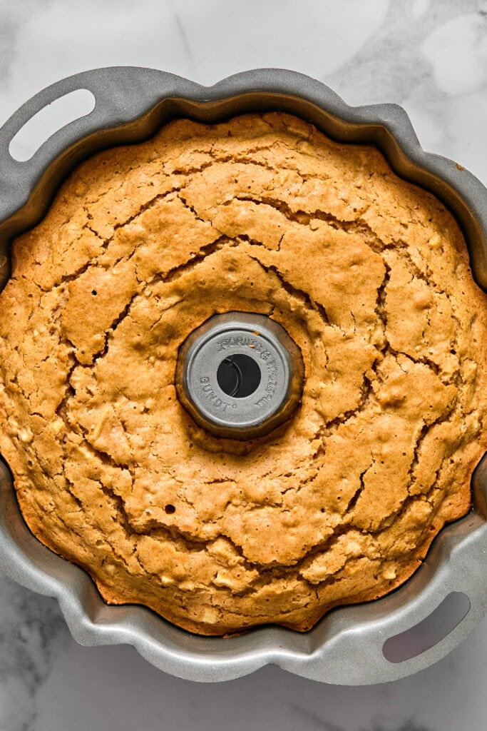 A golden brown vegan gluten-free apple cake with a cracked top sits in a metal Bundt pan on a marble surface, viewed from above.