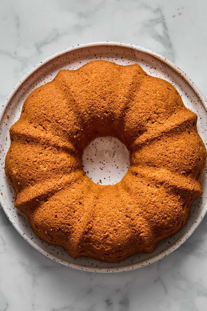 A golden-brown vegan gluten-free apple cake sits on a white speckled plate, viewed from above, with a marble surface in the background.