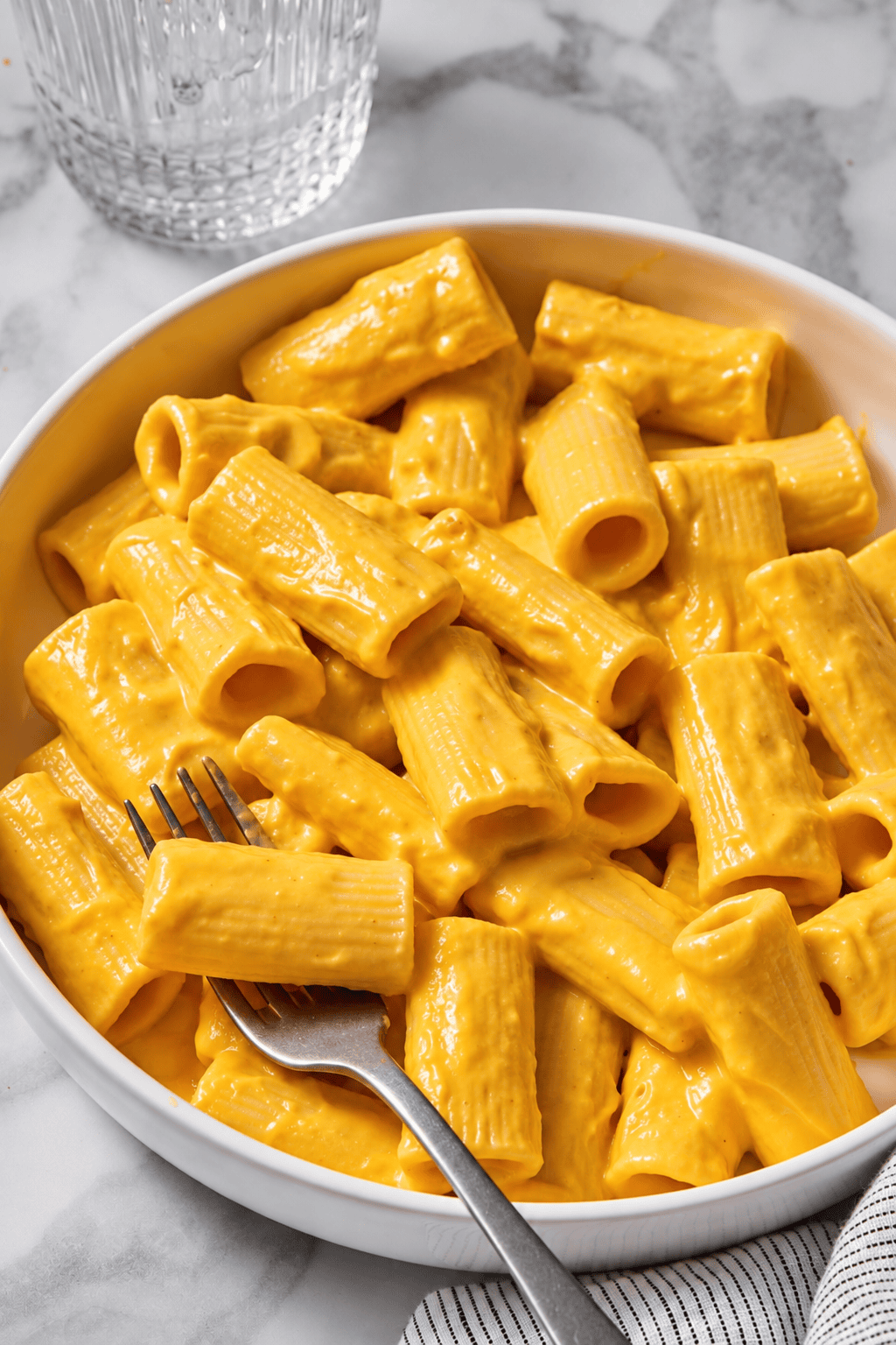 A white bowl filled with rigatoni pasta coated in a creamy orange cheese sauce, inspired by Creamy Vegan Pumpkin Garlic Pasta, with a fork resting on the side. A glass of water and a striped napkin are nearby on a marble surface.