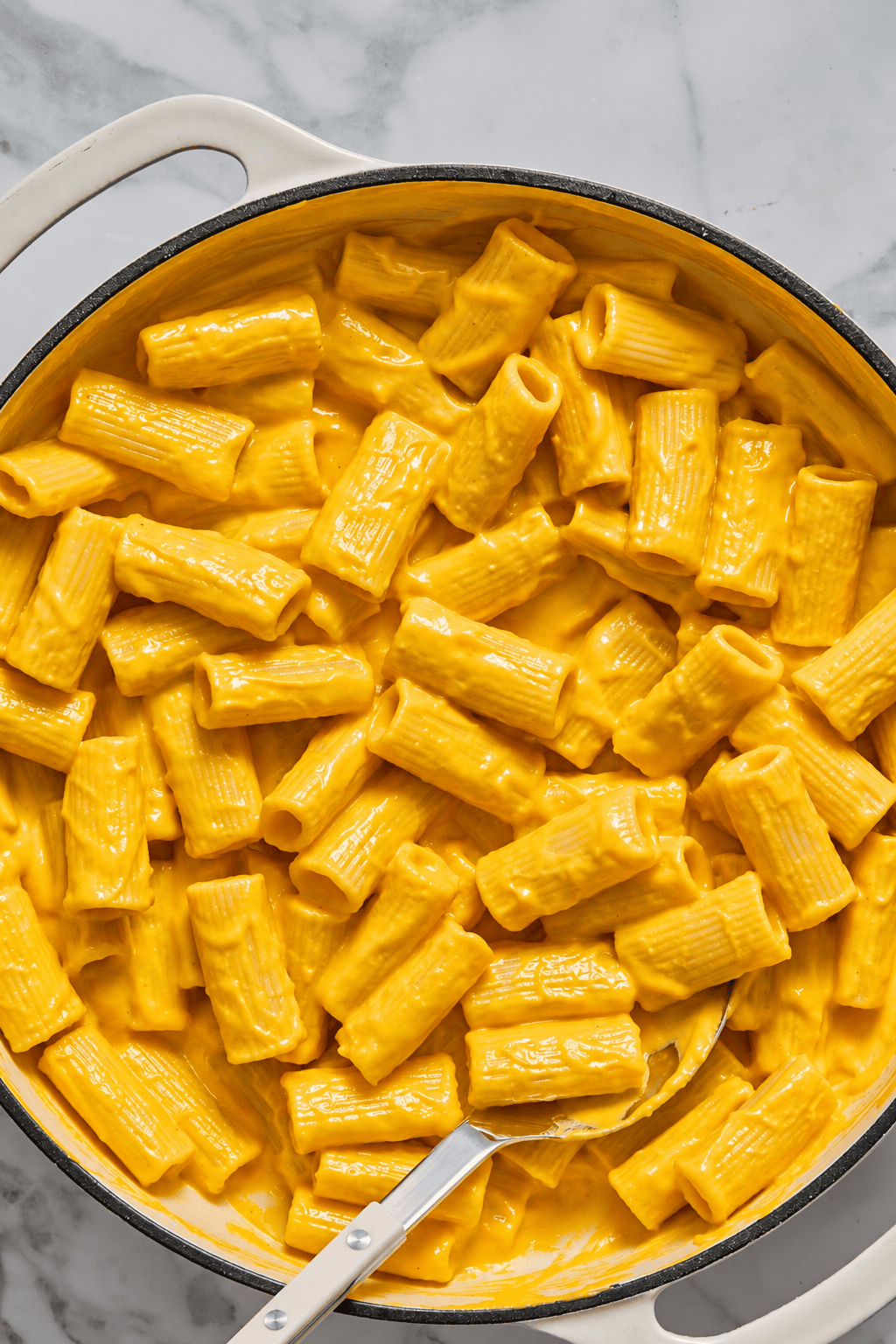A pot filled with rigatoni pasta coated in creamy, bright orange cheese sauce sits on a marble surface. A metal spoon rests in the pot, ready to serve this irresistible Creamy Vegan Pumpkin Garlic Pasta.