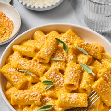A bowl of Creamy Vegan Pumpkin Garlic Pasta in a luscious orange sauce, topped with grated cheese and fresh sage leaves, sits on a table with a fork, a glass of water, and small dishes of cheese and crumbs nearby.