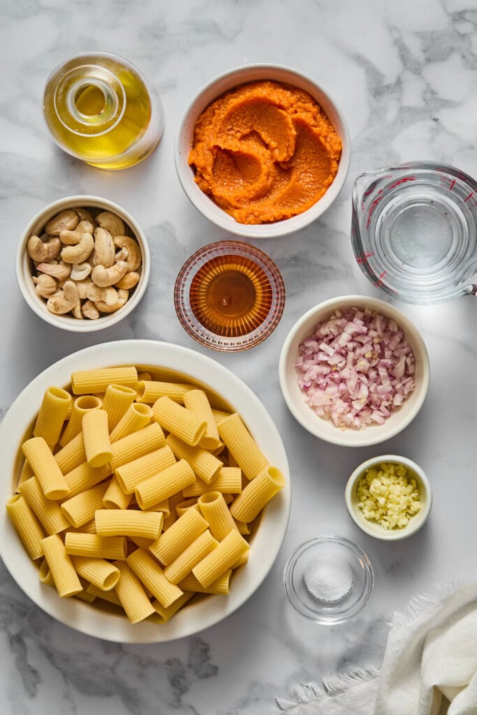 Uncooked rigatoni pasta, cashews, chopped onion, minced garlic, a cup of water, olive oil, a bowl of orange sauce for creamy vegan pumpkin garlic pasta, and a small bowl of amber liquid rest on a marble countertop.