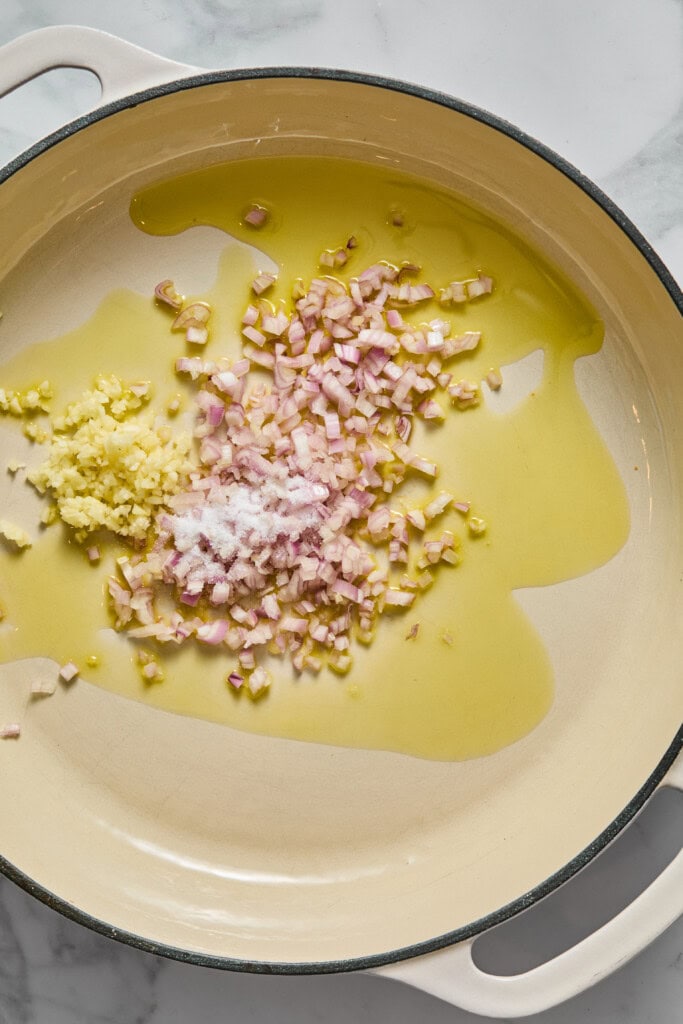 Chopped shallots, minced garlic, and salt in olive oil sizzle in a large cream-colored pan on a marble counter, ready to be transformed into a delicious Creamy Vegan Pumpkin Garlic Pasta.