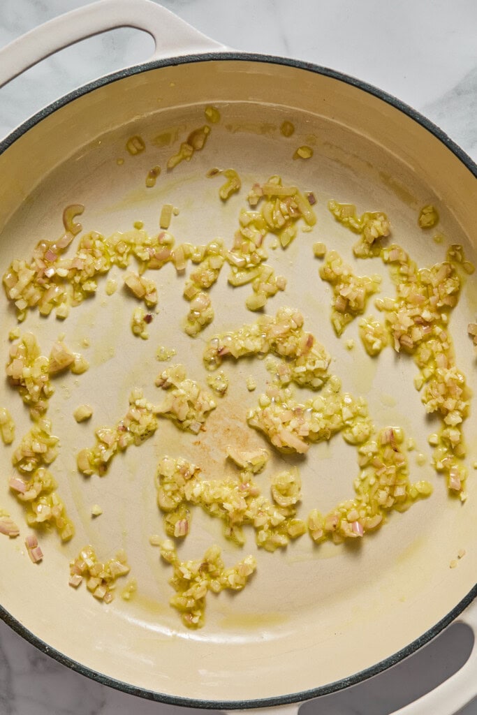 Diced onions and garlic sautéing in oil in a large cream-colored pan on a marble countertop, forming the aromatic base for a creamy vegan pumpkin garlic pasta.