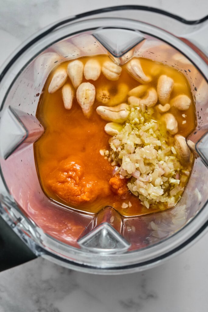Top view of a blender containing cashews, chopped garlic, ginger, a reddish-orange paste, and a brown liquid—ingredients for a creamy Vegan Pumpkin Garlic Pasta sauce—ready to be blended, on a light marble surface.