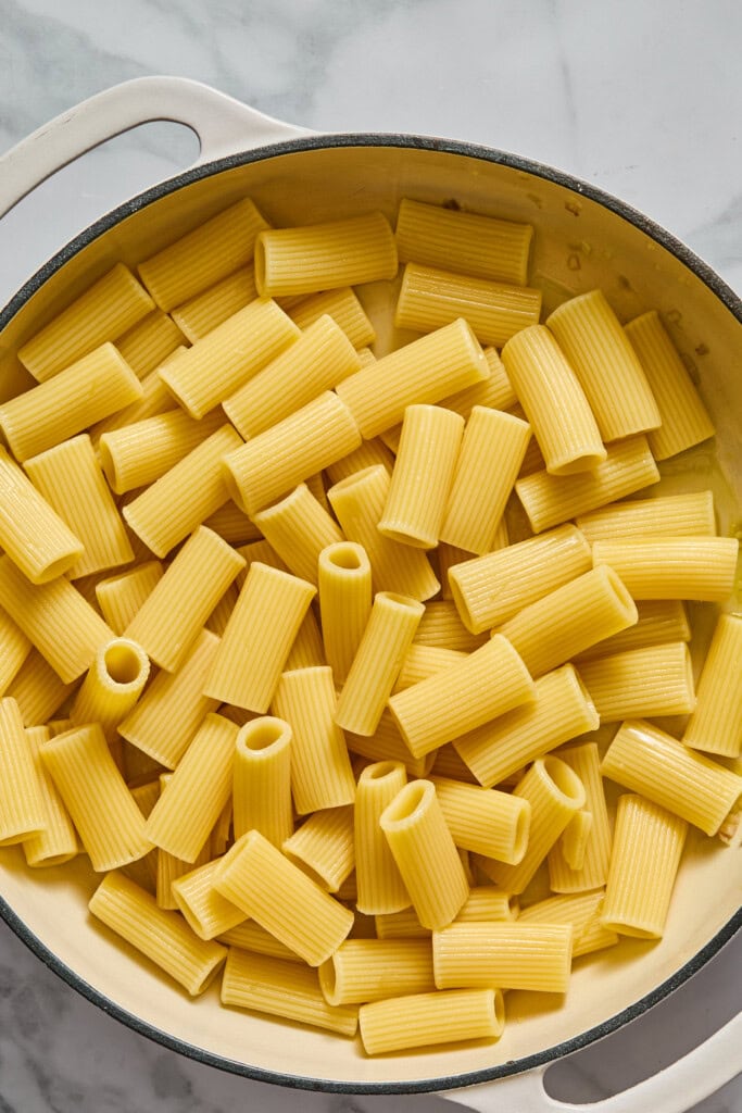 A large pot filled with cooked rigatoni pasta sits on a marble surface, ready to be transformed into a luscious Creamy Vegan Pumpkin Garlic Pasta. The pale yellow, tubular pieces with ridges are arranged in a single layer, awaiting the rich pumpkin and garlic sauce.