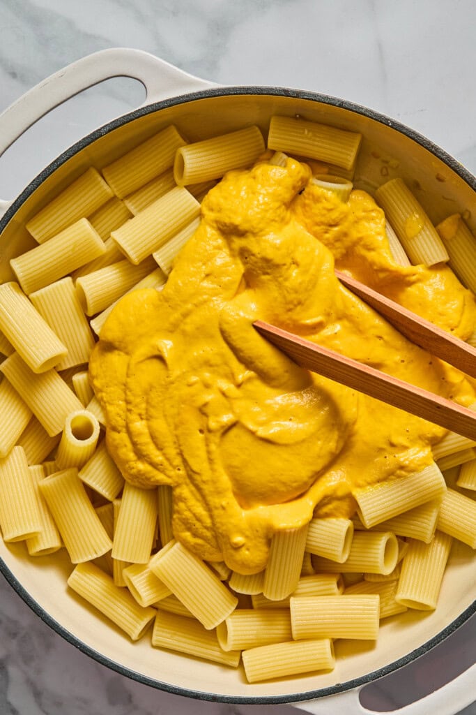 A pot of rigatoni pasta with creamy yellow sauce, reminiscent of Creamy Vegan Pumpkin Garlic Pasta, being mixed with wooden utensils on a marble countertop.