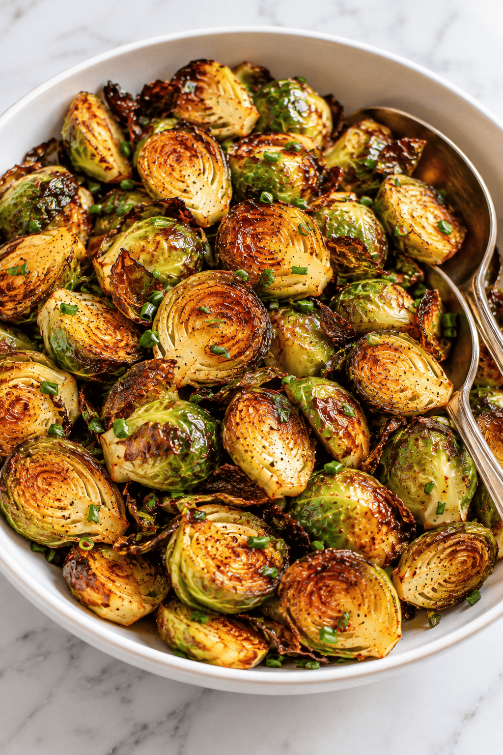 A white bowl filled with maple Brussels sprouts, garnished with chopped herbs and served with two metal serving spoons on a marble surface.