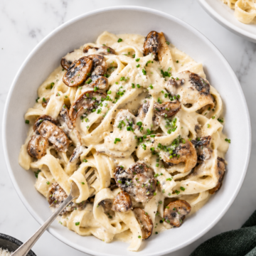 A bowl of creamy vegan mushroom pasta with sautéed mushrooms, garnished with chopped chives. A fork rests in the bowl, and small dishes of chives and grated cheese are nearby on a marble surface.