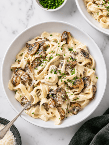 A bowl of creamy vegan mushroom pasta with saut&eacute;ed mushrooms, garnished with chopped chives. A fork rests in the bowl, and small dishes of chives and grated cheese are nearby on a marble surface.