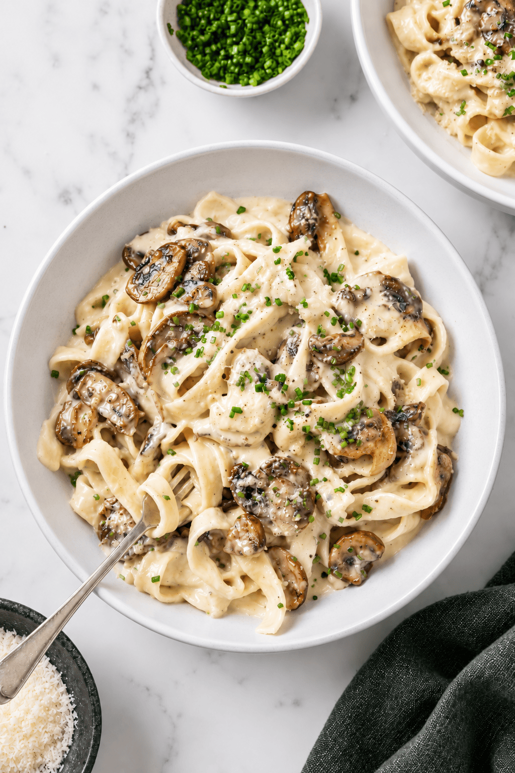 A bowl of creamy vegan mushroom pasta with sautéed mushrooms, garnished with chopped chives. A fork rests in the bowl, and small dishes of chives and grated cheese are nearby on a marble surface.