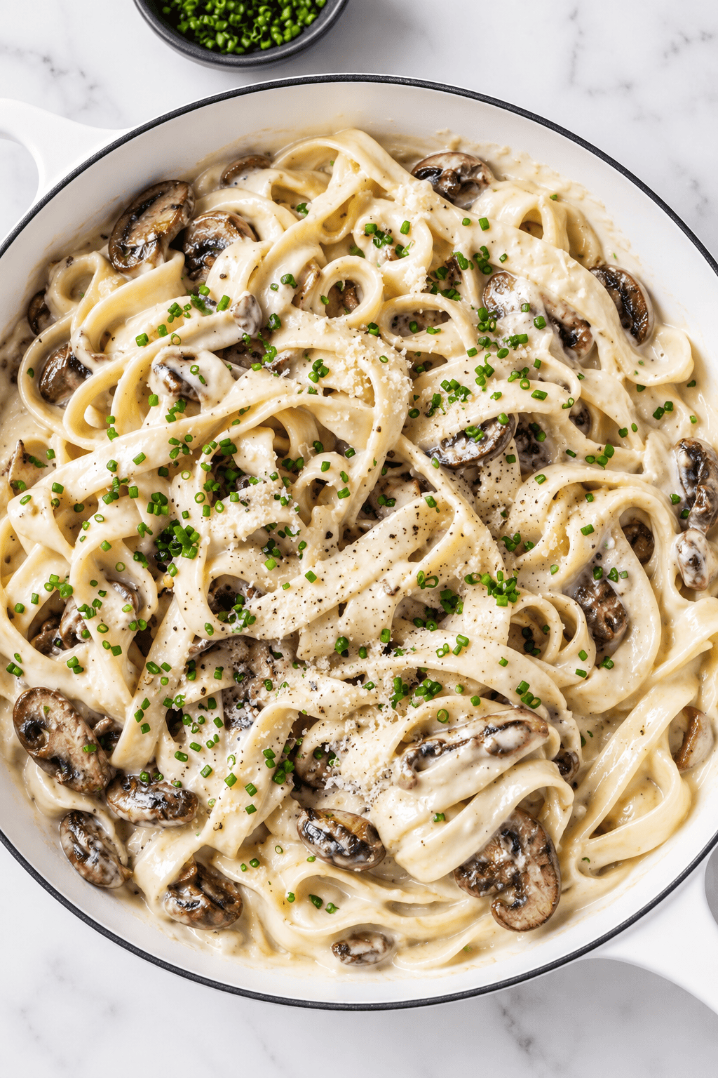 A pan of creamy vegan mushroom pasta with sautéed mushrooms, topped with chopped chives and black pepper, on a white marble surface.