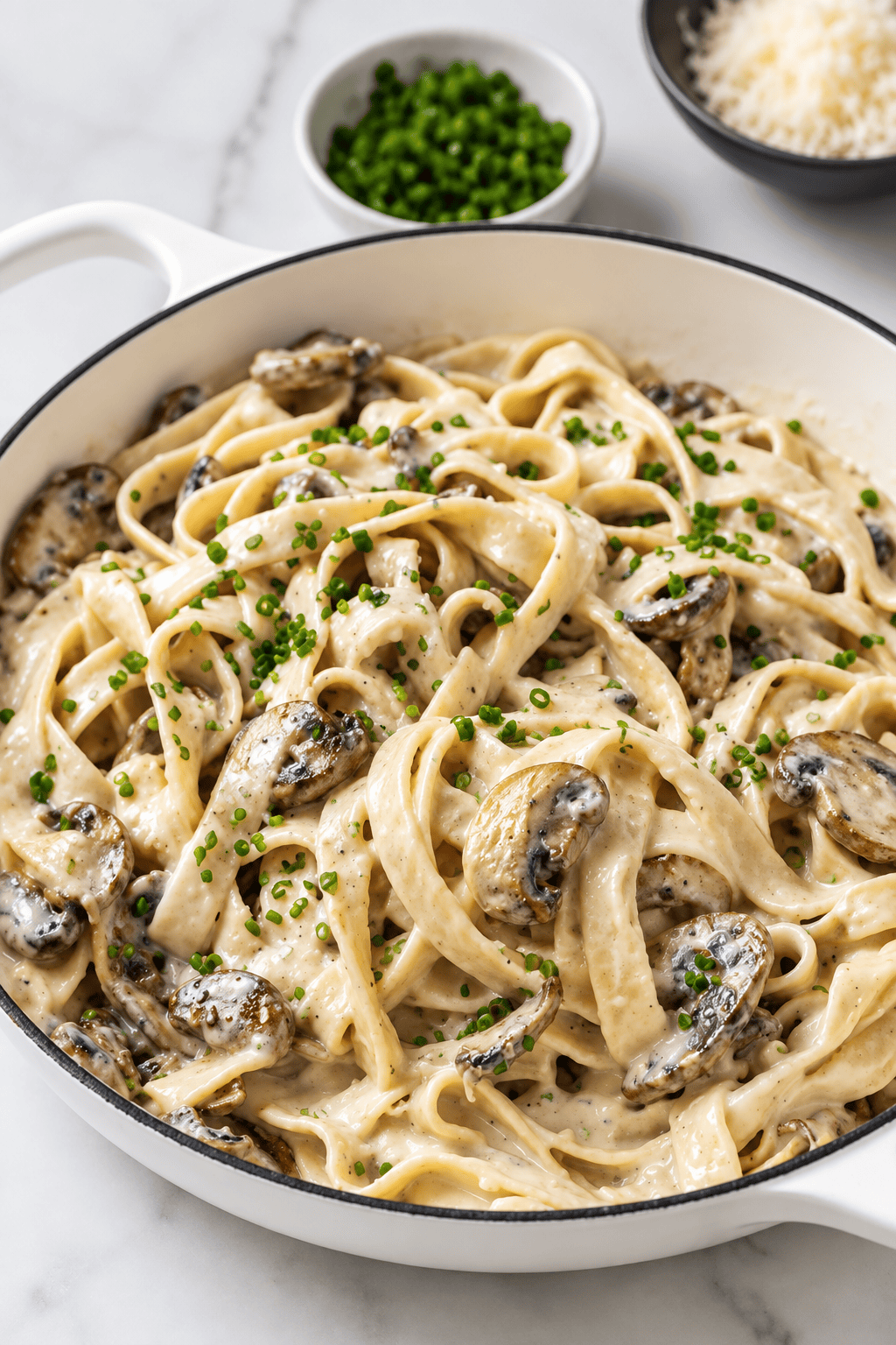 Creamy Vegan Mushroom Pasta features fettuccine tossed in a luscious, dairy-free mushroom sauce and topped with fresh chopped chives, served in a white dish. Small bowls of grated vegan cheese and extra chives sit invitingly in the background.