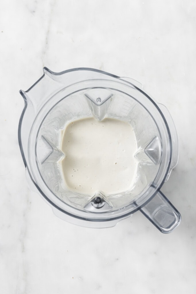 A top-down view of a blender containing a small amount of creamy white liquid on a light marble surface, perfect for preparing the luscious sauce base for Creamy Vegan Garlic Mushroom Fettuccini.