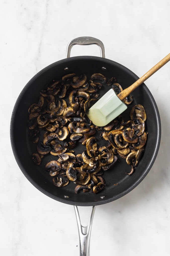 Sliced mushrooms being sautéed in a black pan with a green spatula on a white surface, perfect for preparing Creamy Vegan Garlic Mushroom Fettuccini.