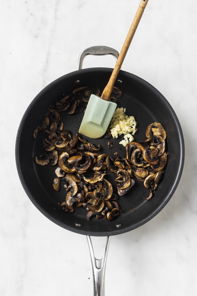 A black skillet with cooked sliced mushrooms and a small pile of minced garlic, stirred with a green spatula, sits on a white marble surface—perfectly capturing the savory beginnings of a Creamy Vegan Garlic Mushroom Fettuccini.