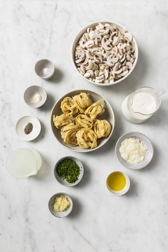 Bowls of sliced mushrooms, uncooked tagliatelle, cream, grated cheese, chopped parsley, olive oil, minced garlic, pepper, salt, and a cup of broth are arranged on a white marble surface—everything you need to prepare a delicious Creamy Vegan Garlic Mushroom Fettuccini.