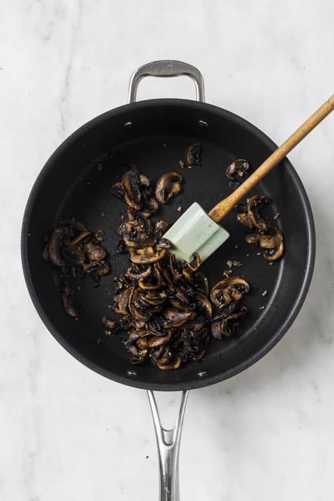 A black skillet with sautéed sliced mushrooms for Creamy Vegan Garlic Mushroom Fettuccini and a light green spatula on a white marble surface.