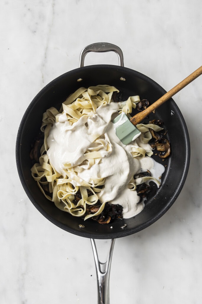 A black pan with cooked pasta, sautéed mushrooms, and creamy white sauce being stirred with a spatula on a marble surface—perfect for serving up Creamy Vegan Garlic Mushroom Fettuccini.