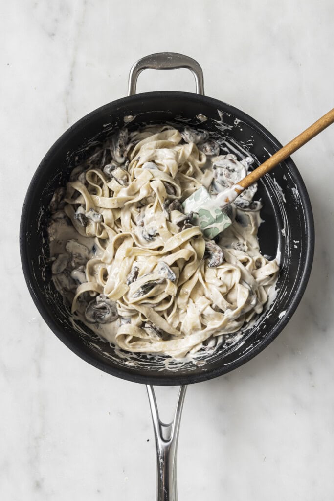 A black skillet filled with Creamy Vegan Garlic Mushroom Fettuccini sits on a marble surface. A spatula with a wooden handle rests in the pan, gently stirring the creamy pasta and mushrooms.