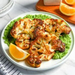 A plate with roasted cauliflower steaks on a bed of green sauce, garnished with lemon wedges. A striped napkin, cutlery, lemon slices, a white oil bottle, and stacked plates rest nearby on a white marble surface.