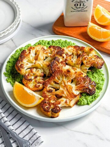 A plate with roasted cauliflower steaks on a bed of green sauce, garnished with lemon wedges. A striped napkin, cutlery, lemon slices, a white oil bottle, and stacked plates rest nearby on a white marble surface.