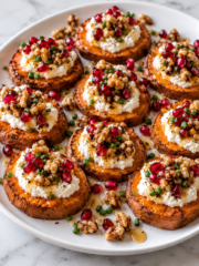 Sweet Potato Rounds with Vegan Ricotta are topped with chopped walnuts, pomegranate seeds, honey, chives, and black pepper on a white plate, arranged neatly on a marble surface.