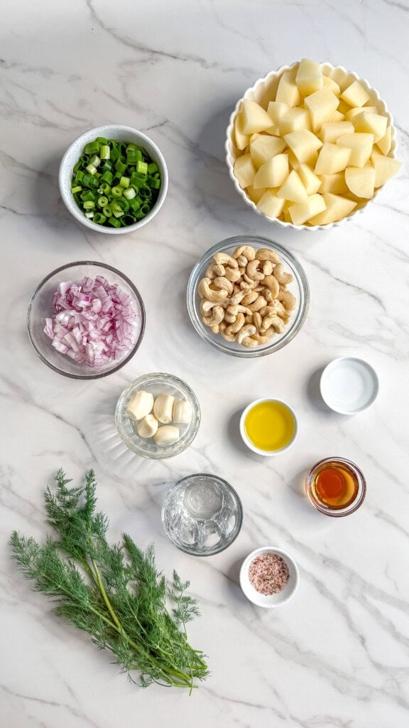 A top-down view of neatly arranged ingredients on a marble surface for Vegan Potato Salad, including chopped potatoes, sliced green onions, diced red onion, raw cashews, garlic cloves, olive oil, vinegar, salt, fresh dill, and a bowl of amber liquid.
