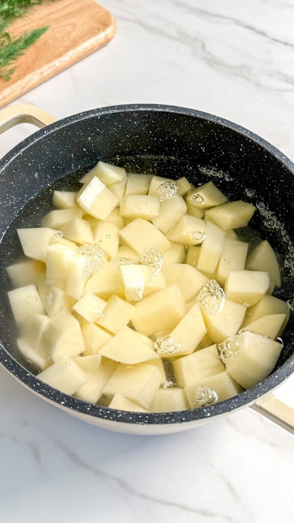 A black pot filled with water and cubed potatoes sits on a white marble countertop, ready for boiling—a fresh start for a Vegan Potato Salad. A wooden cutting board with herbs is visible in the background.