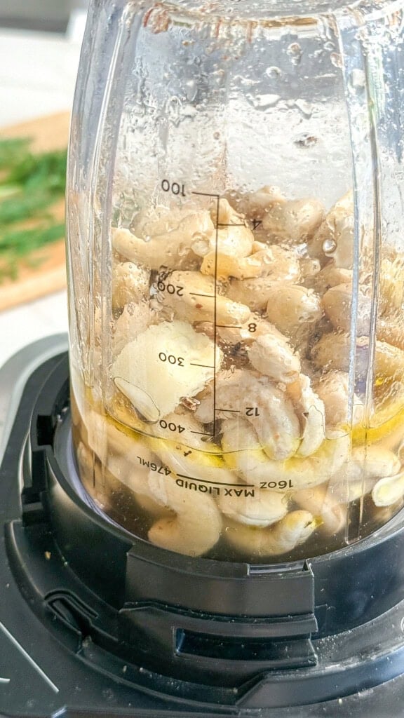 Close-up of a blender cup filled with raw cashews, garlic, and olive oil for Vegan Tzatziki. Measurement marks are visible on the slightly wet cup, showing ingredients ready to be blended into a creamy dressing for Vegan Potato Salad.