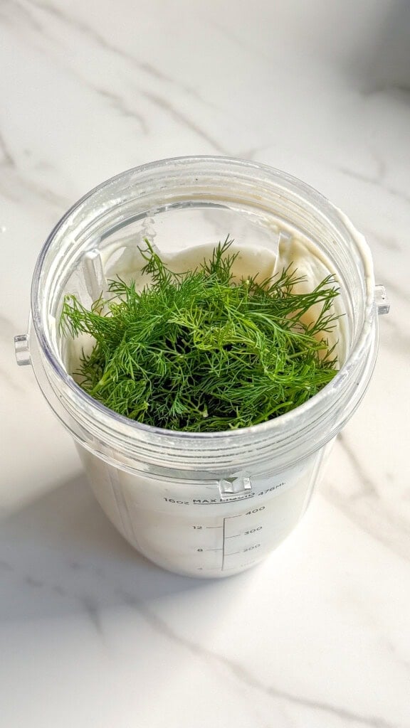 A blender cup filled with creamy white yogurt and topped with a bunch of fresh green dill, perfect for making Vegan Tzatziki or adding a tangy twist to your favorite potato salad, sits on a white marble countertop.