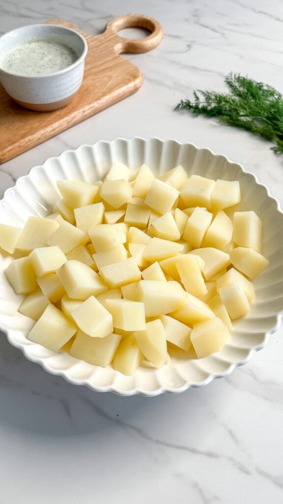A white scalloped bowl filled with peeled and diced potatoes sits on a marble surface. In the background are a small bowl of creamy vegan tzatziki sauce on a wooden board and fresh dill, perfect for assembling a refreshing potato salad.