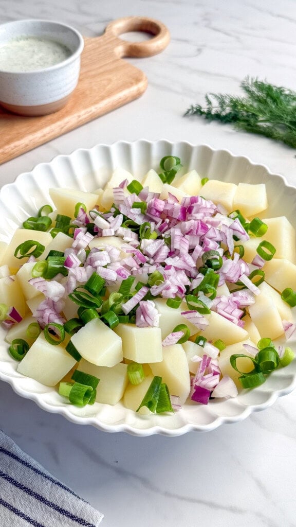 A white bowl filled with chopped boiled potatoes, topped with diced red onions and sliced green onions, sits on a marble surface. A bowl of Vegan Tzatziki dressing and fresh dill are in the background, perfect for this refreshing Vegan Potato Salad.