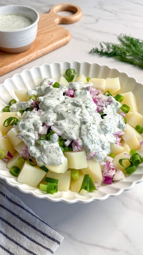 A white scalloped bowl with cubed potatoes, chopped red onion, and sliced green onions, topped with a creamy Vegan Tzatziki dill sauce. A bowl of dressing, fresh dill, and a striped napkin are nearby on a marble surface.