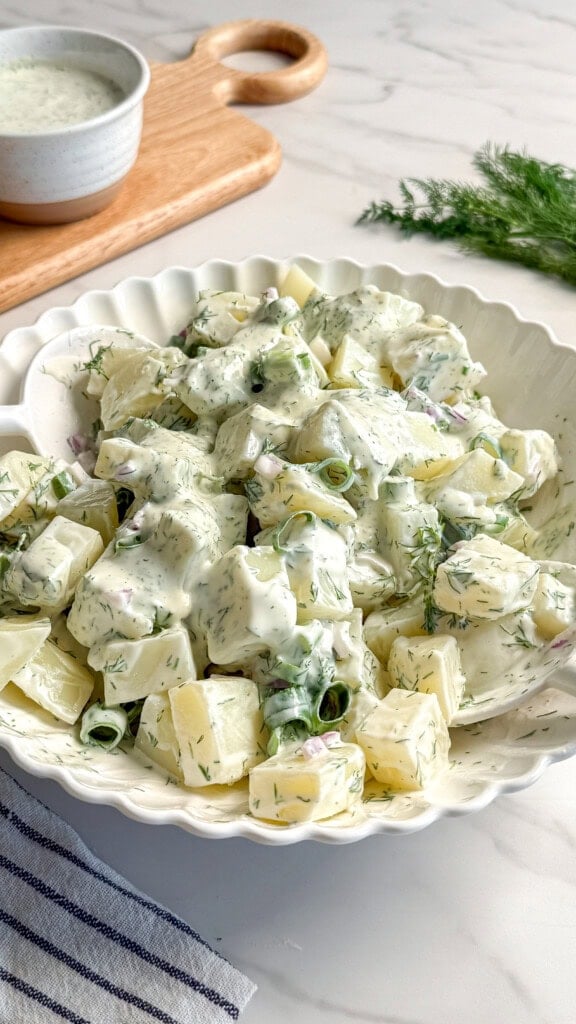 A white bowl filled with creamy vegan potato salad garnished with green herbs sits on a marble countertop. A small bowl of vegan tzatziki and a sprig of fresh dill are in the background.