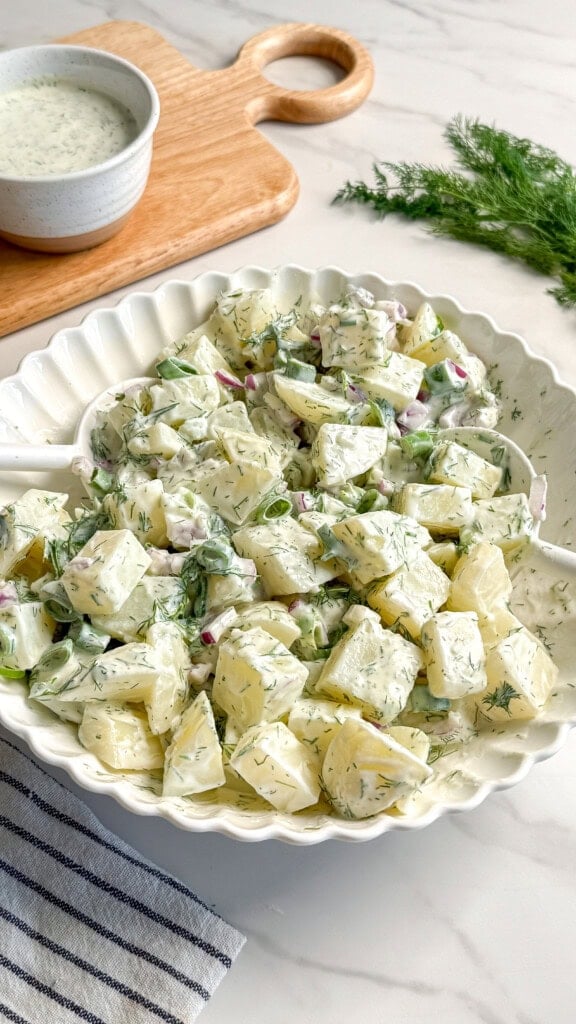A white bowl filled with creamy Vegan Potato Salad garnished with fresh dill, with a striped napkin, a small bowl of Vegan Tzatziki dressing, and sprigs of dill on a light marble surface in the background.