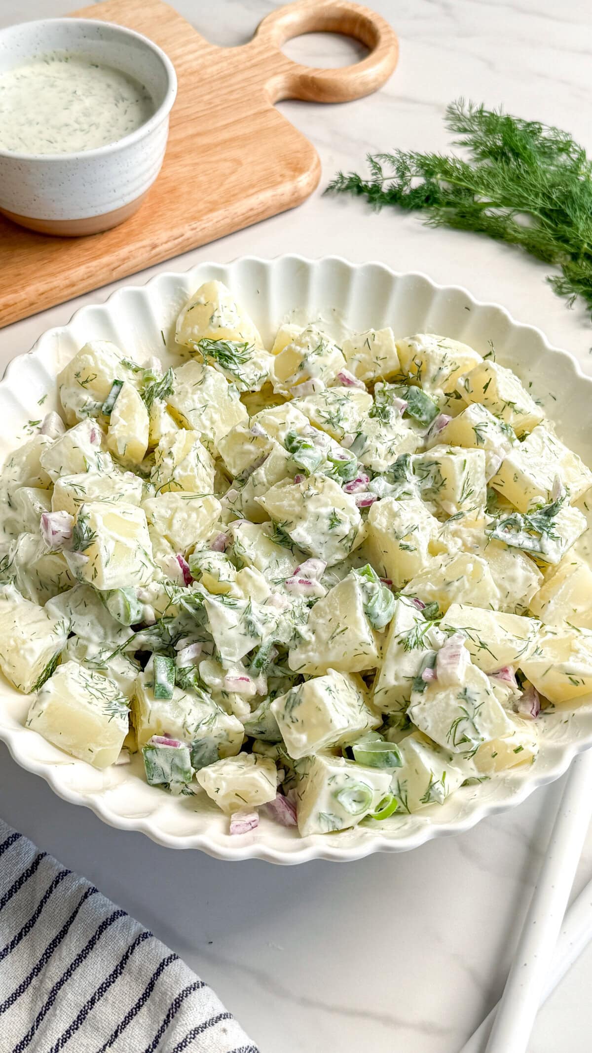 A white scalloped bowl filled with creamy Vegan Potato Salad garnished with fresh dill, with diced red onion and green vegetables visible. A cup of dressing and sprigs of dill rest on a wooden board in the background.